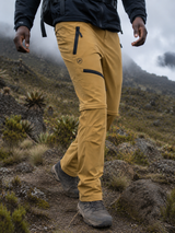 A person hiking outdoors on a rocky mountain path, wearing tan waterproof pants with black zippers and a black jacket.