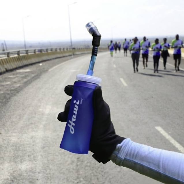 A close-up of a hand wearing a black glove holding a blue hydration bottle with a straw on a road, with a group of runners visible in the background on a bridge.
