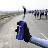 A close-up of a hand wearing a black glove holding a blue hydration bottle with a straw on a road, with a group of runners visible in the background on a bridge.