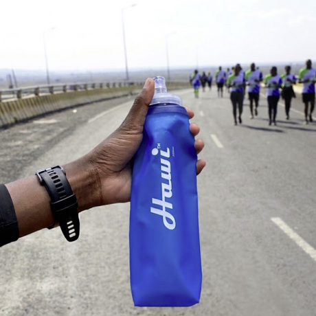A close-up of a hand holding a blue Hydration Pro water bottle with a clear cap, with a road and running group in the background.