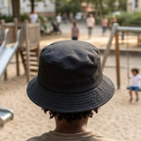 A child wearing a black bucket hat, brown t-shirt, and denim shorts sits on a wooden playground staircase, looking down at a sandy playground with children and trees in the background.