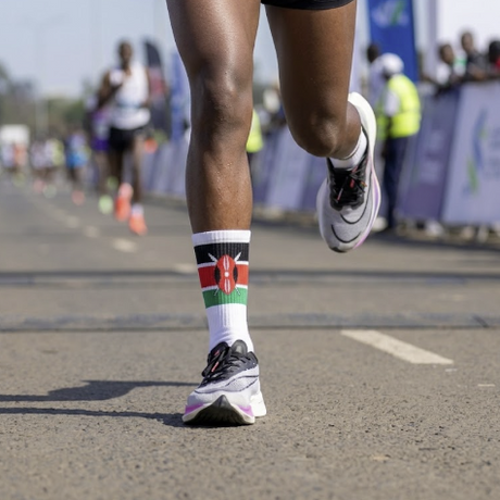 A runner's lower legs in black athletic shorts, white running socks with a Kenyan flag design, and white running shoes on an asphalt road during a race.