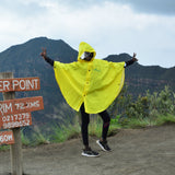 A person wearing a bright yellow rain poncho stands with arms outstretched in front of mountain peaks under a cloudy sky, next to a wooden sign reading 'Oloonongot Crater Point' and other location details.