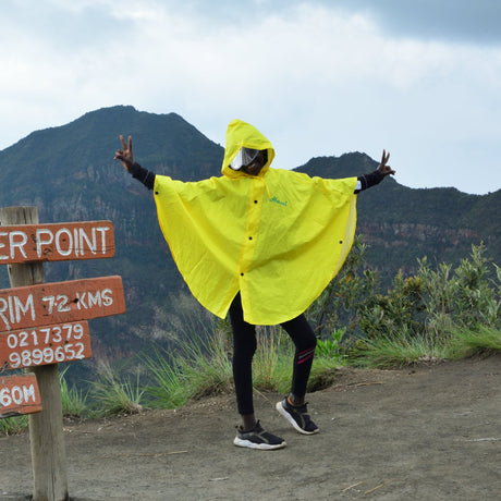 A person wearing a bright yellow rain poncho stands with arms outstretched in front of mountain peaks under a cloudy sky, next to a wooden sign reading 'Oloonongot Crater Point' and other location details.