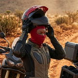 Two motorcyclists pause on a dusty dirt road in a desert setting. The rider on the left adjusts a red and black helmet with a red balaclava, while the rider on the right wipes a black motorcycle with a blue cloth.