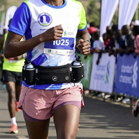 A female marathon runner wearing a blue, white, and green athletic jersey, pink shorts, and a black hydration belt running on a road during a race.