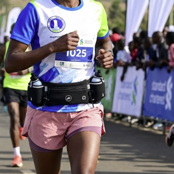A female marathon runner wearing a blue, white, and green athletic jersey, pink shorts, and a black hydration belt running on a road during a race.