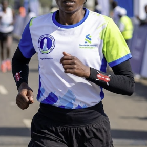 A female runner wearing a white, blue, and lime green athletic jersey with black long sleeves, black running shorts, and black-and-white socks, captured mid-stride on an outdoor road race.