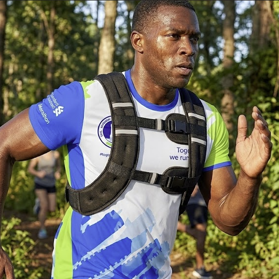 A male runner wearing a white, blue, and green athletic jersey with a black hydration vest is running through a wooded trail.