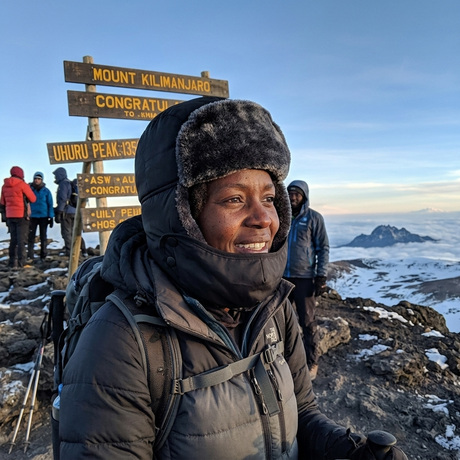 A smiling woman wearing a dark winter puffer jacket and a fuzzy dark hat stands on a mountain peak with a signpost reading Mount Kilimanjaro and Uhuru Peak in the background, with snow and distant mountains under a blue sky.