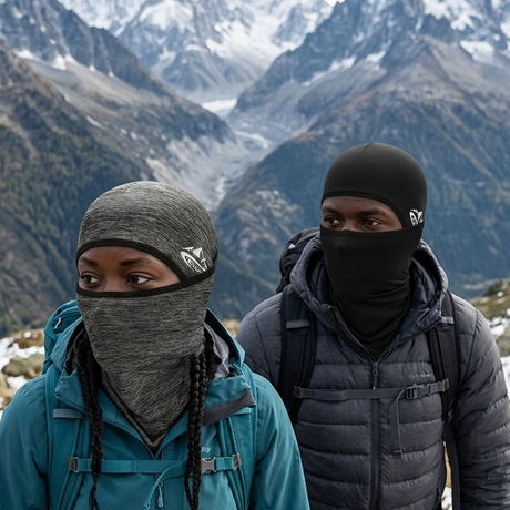 Two hikers wearing ski balaclavas and outdoor gear standing on a snowy mountain ridge with a dramatic alpine valley and snow-capped peaks in the background.